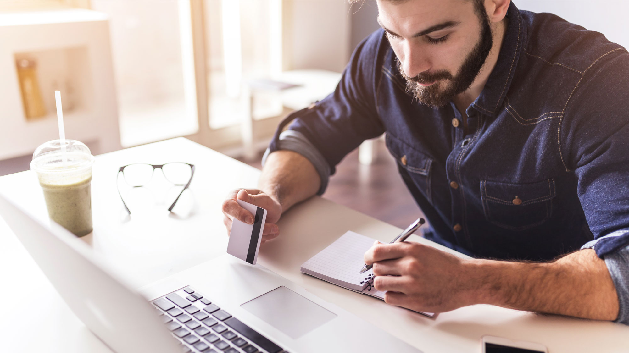 Man with credit card in one hand, computer in front of him and a writing in a notebook